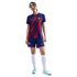 A woman wears an FC Barcelona soccer kit with a blue and red striped jersey, blue shorts, white socks, and light blue cleats, standing against a black background.