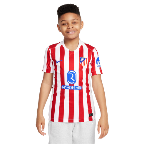 A smiling young boy wearing a red and white striped Atlético Madrid soccer jersey and light grey shorts, standing against a plain white background.