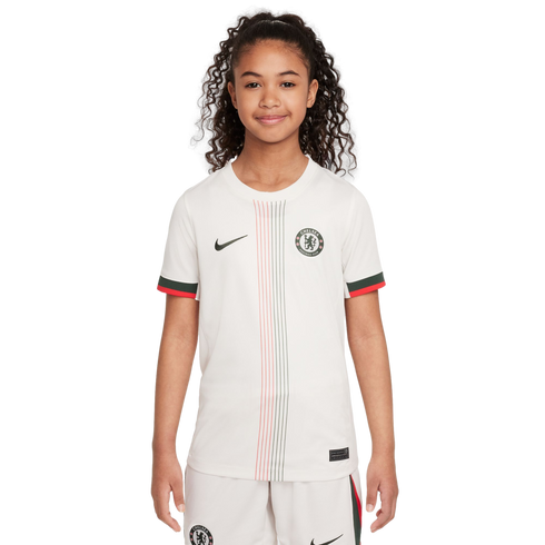 A young girl with curly hair posing in a white Chelsea FC soccer jersey with red and green accents and matching shorts.