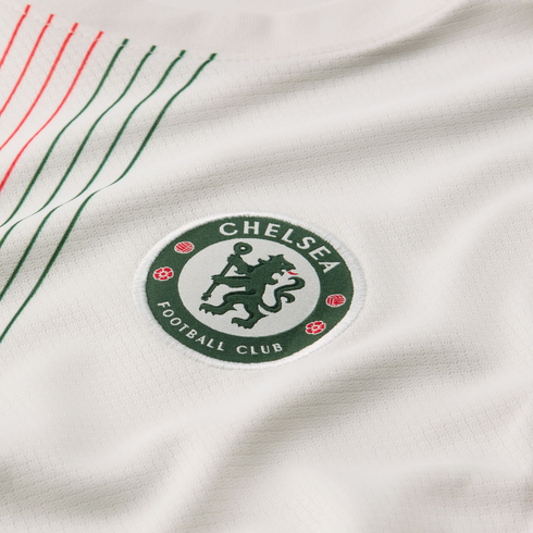 Close-up of a green Chelsea Football Club crest on a white textured jersey with thin red and green diagonal stripes in the corner.