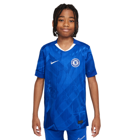 A young boy with braided hair smiling while wearing a blue Nike Chelsea FC football jersey against a plain white background.