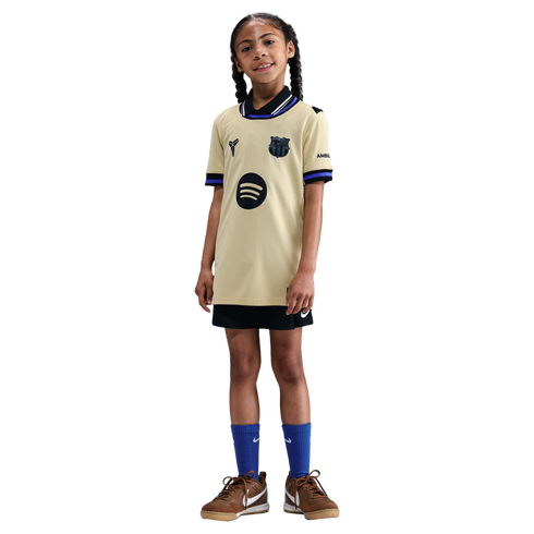 Young girl posing in a gold FC Barcelona Mamba jersey, black shorts, blue socks, and brown sneakers against a black background.