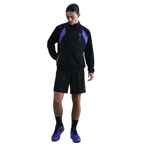 A man wearing a black tracksuit jacket with purple shoulder panels, black shorts, and purple sneakers against a solid black background.