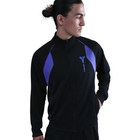 Profile of a person with long hair tied back wearing a black zip-up jacket with purple panels and a logo on the chest, set against a solid black background.