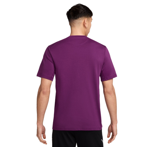 Rear view of a man wearing a plain purple short-sleeved t-shirt and black shorts against a solid black background.