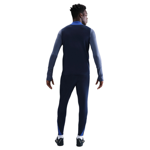 Rear view of a man wearing a navy blue and light blue long-sleeved athletic top and matching pants with black soccer cleats against a white background.