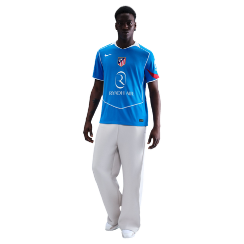 Man in a blue Atletico Madrid soccer jersey and white wide-leg trousers against a black background.