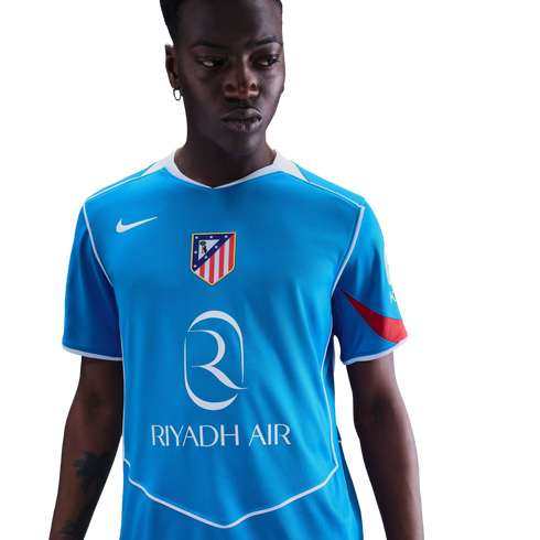 A man with a serious expression wears a light blue Nike soccer jersey with the Atletico Madrid crest and Riyadh Air logo, set against a solid black background.