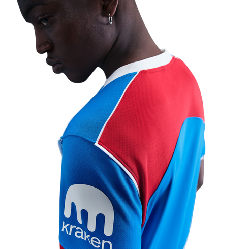 A person wearing a blue and red athletic jersey with a white 'Kraken' logo on the sleeve, looking down against a black background.