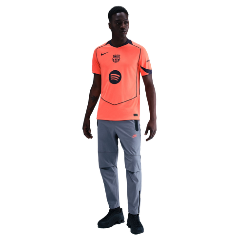 A man modeling a bright orange Nike FC Barcelona jersey, grey athletic pants, and black sneakers against a black background.