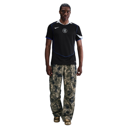 Man wearing a black Nike Chelsea FC jersey, camouflage cargo pants, and gold metallic sneakers against a black background.