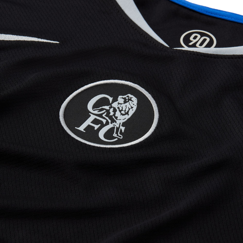 Close-up of a black Chelsea FC jersey with a white embroidered circular lion crest and "CFC" lettering. The collar features blue and white trim and a "90" logo inside.