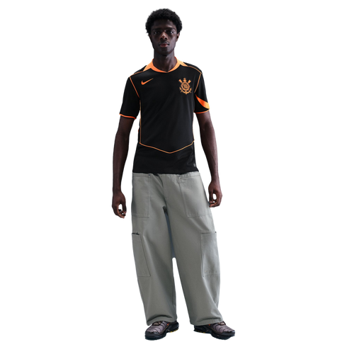 Man wearing a black Nike soccer jersey with orange trim, baggy grey cargo pants, and sneakers, standing against a solid black background.