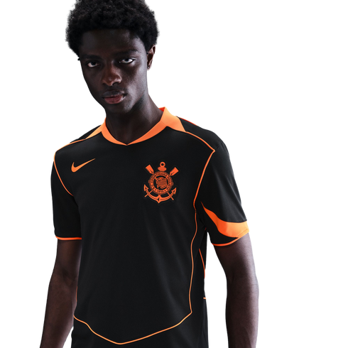 A Black man wearing a black soccer jersey with bright orange trim and a Corinthians crest against a black background.
