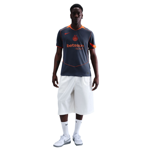 A man wearing a grey and orange Inter Milan training jersey, white knee-length shorts, and white sneakers, standing against a solid black background.