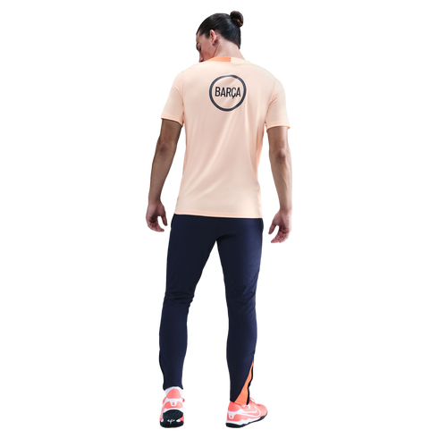 Rear view of a man wearing a peach "BARÇA" t-shirt, navy joggers, and orange sneakers, set against a black background.