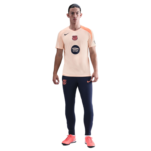 A full-length photo of a man wearing a peach FC Barcelona Nike training shirt, navy blue joggers, and coral soccer shoes against a black background.