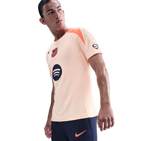 A man wearing a peach Nike FC Barcelona training top and navy shorts, looking off-camera against a black background.