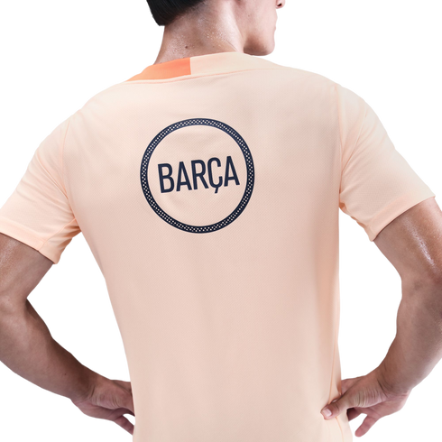 Back view of a person wearing a light orange t-shirt with a navy blue circular logo reading "BARÇA" on the back.