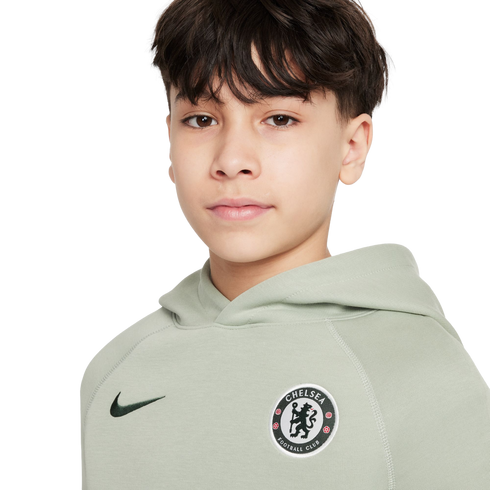 Young person with dark hair wearing a sage green Nike Chelsea Football Club hoodie against a plain white background.