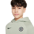 Young person with dark hair wearing a sage green Nike Chelsea Football Club hoodie against a plain white background.
