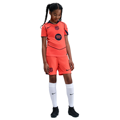 A young girl in a bright orange FC Barcelona soccer kit, white socks, and black cleats, standing against a solid black background.