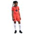 A young girl in a bright orange FC Barcelona soccer kit, white socks, and black cleats, standing against a solid black background.