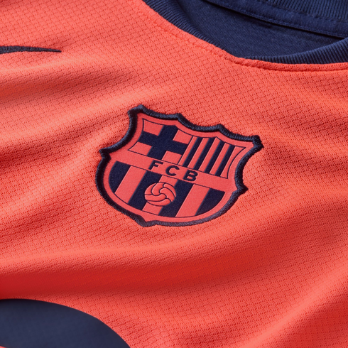 Close-up of a salmon-colored FC Barcelona jersey featuring the embroidered club crest. The shirt has a textured mesh fabric and navy blue collar trim.