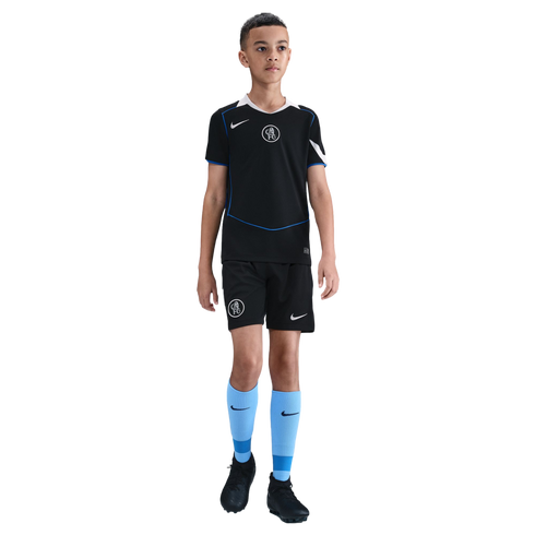 A young boy stands in a black Nike Chelsea FC soccer kit with light blue socks and black cleats, isolated on a black background.