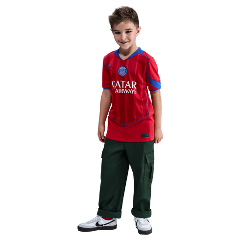 A young boy stands smiling, wearing a red Paris Saint-Germain football jersey, dark green cargo pants, and white sneakers against a black background.