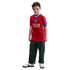 A young boy stands smiling, wearing a red Paris Saint-Germain football jersey, dark green cargo pants, and white sneakers against a black background.