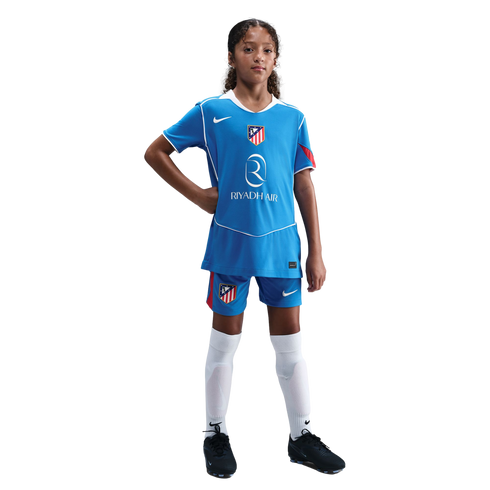 A young girl in a blue Atletico Madrid soccer kit with white socks and black cleats, posing with a hand on her hip against a black background.