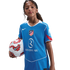 A young girl with curly hair poses in a blue Atlético Madrid soccer jersey, holding a soccer ball under her arm against a black background.