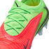 A close-up, high-angle view of a vibrant neon green and bright pink Nike soccer cleat against a black background. The shoe features a textured, knitted upper with matching neon green laces and multiple black Nike swoosh logos. Clear, translucent studs are