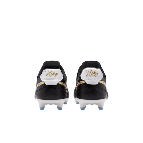 Rear view of black Nike soccer cleats with gold cursive branding on white heel tabs and white soleplates with blue studs against a black background.