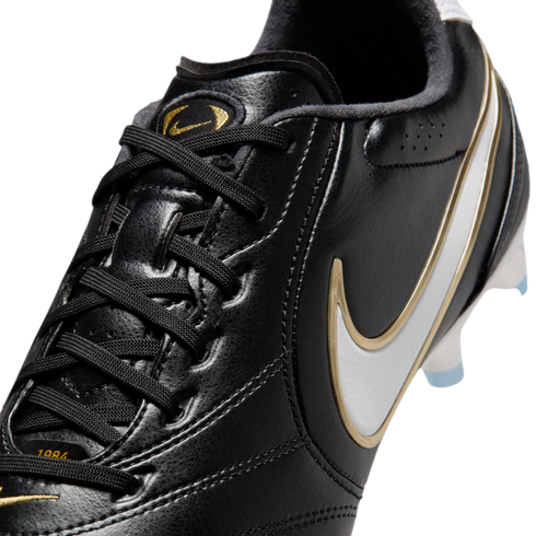 Close-up of a black leather Nike soccer cleat with black laces, gold branding on the tongue, and a white Nike swoosh with a gold border on the side.