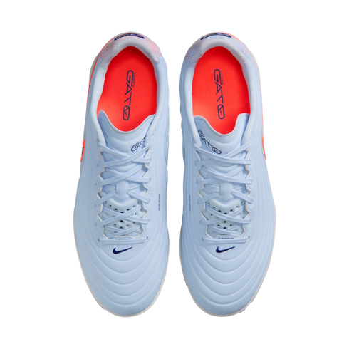 A top-down view of a pair of light blue Nike Tiempo Gato indoor soccer shoes with bright orange insoles, set against a solid black background.