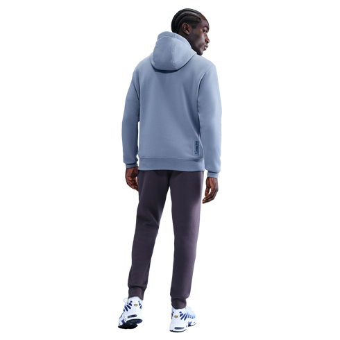 Rear view of a man with braids wearing a light blue hoodie and dark grey joggers against a black background.