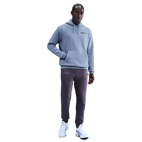 A man models a light blue Nike Spurs hoodie, dark purple sweatpants, and white sneakers against a black background.