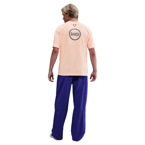 Back view of a man wearing a peach-colored Nike Barça t-shirt and baggy blue pants against a black background.