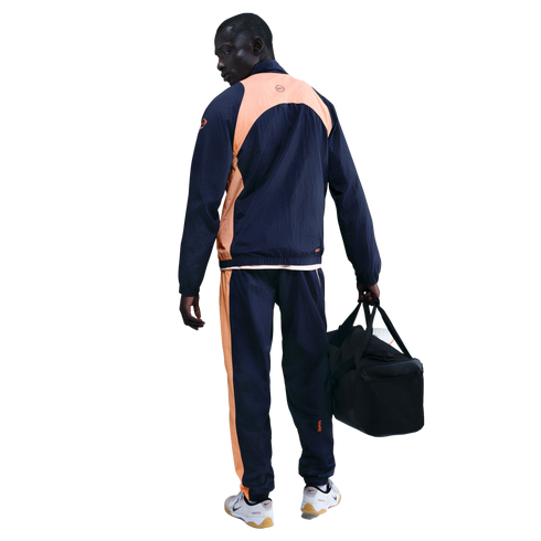 A man in a navy blue and peach tracksuit, seen from behind, holding a black gym bag against a black background.