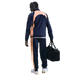 A man in a navy blue and peach tracksuit, seen from behind, holding a black gym bag against a black background.
