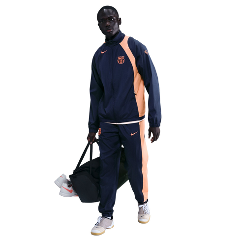 A man wearing a navy and orange Nike FC Barcelona tracksuit carries a black duffel bag against a solid black background.