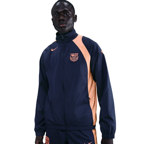 A man wearing a navy blue and peach Nike FC Barcelona windbreaker tracksuit against a black background.