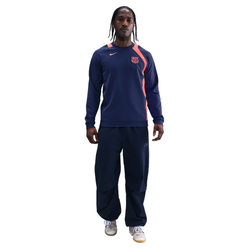 Man in a navy Nike FC Barcelona sweatshirt with orange accents and matching track pants, standing against a black background.
