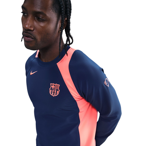 Man with braids wearing a navy and pink Nike FC Barcelona long-sleeve training shirt, looking away against a black background.