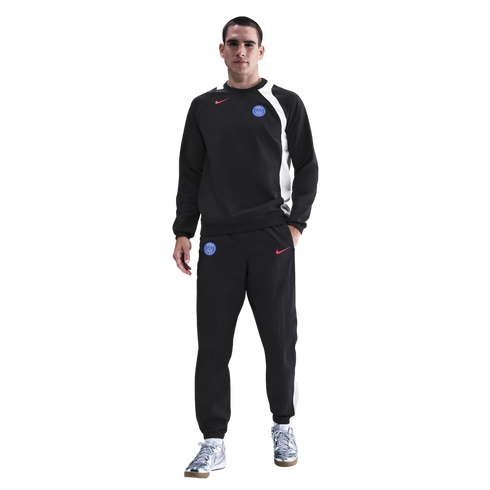 Man wearing a black Nike Paris Saint-Germain tracksuit and silver sneakers against a black background.