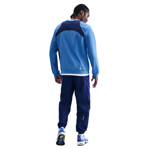 Back view of a man with braided hair wearing a blue and navy athletic tracksuit and sneakers against a solid black background.