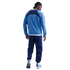 Back view of a man with braided hair wearing a blue and navy athletic tracksuit and sneakers against a solid black background.