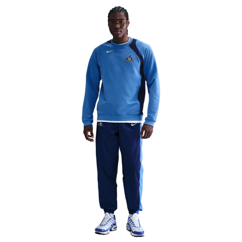 Man wearing a blue Tottenham Hotspur Nike sweatshirt, navy track pants, and blue and white sneakers against a black background.